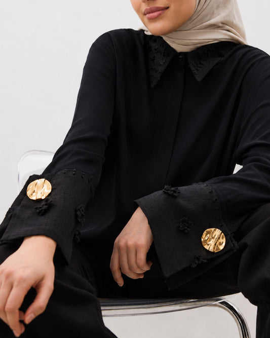 Chemise longue noire élégante avec détails dorés aux poignets, coupe fluide.